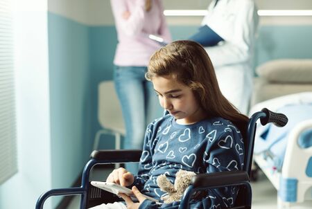 Cute girl in wheelchair at the hospital using a tablet, her mother and the doctor are talking in the backgroundの写真素材