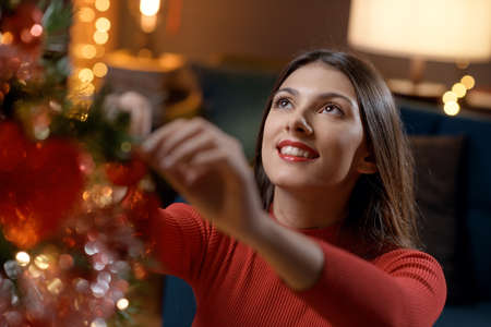 Beautiful woman decorating her Christmas tree at home, she is hanging ornaments and baubles, holidays and celebrations conceptの写真素材