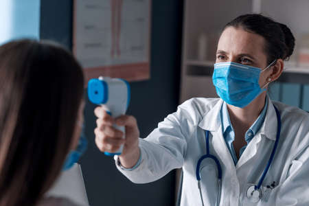 Professional doctor with surgical mask measuring a patient's temperature using an infrared forehead thermometerの写真素材