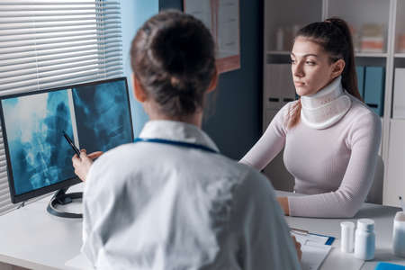 Doctor explaining a x-ray to the patient and pointing at the computer screen, the woman is wearing a cervical collarの写真素材