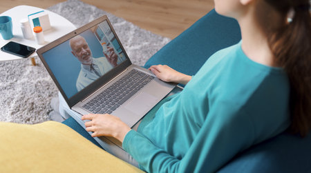 Woman video calling her doctor at home, she is connecting with her laptop and sitting on the sofa, online doctor conceptの写真素材