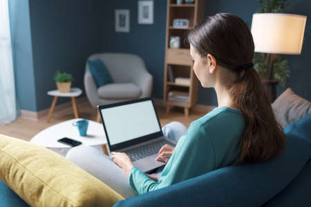 Young woman relaxing at home, she is resting on the sofa and connecting online with her laptop