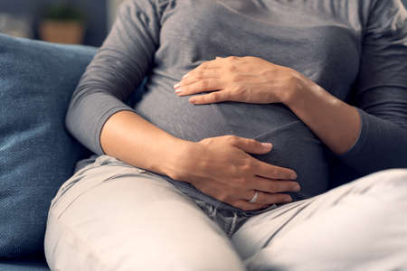 Pregnant woman sitting on the sofa and holding her belly, hands close upの写真素材