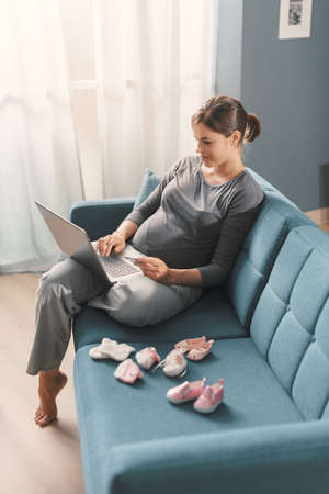 Pregnant woman sitting on the sofa and doing online shopping at home, she is connecting with her...