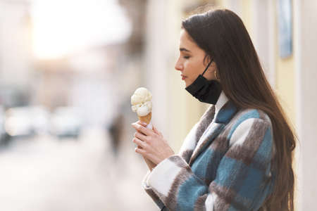 Young happy woman standing in the street and having a delicious ice cream, she is wearing a face maskの写真素材