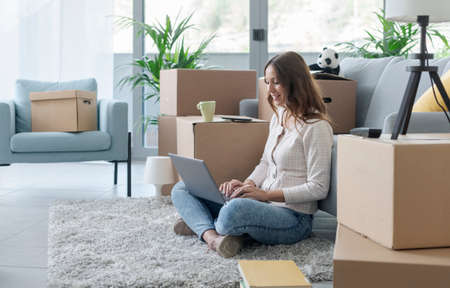 Happy young woman moving in her new apartment, she is sitting on the floor and connecting with her...