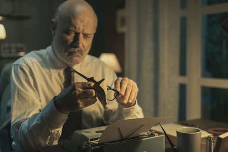Corporate businessman sitting at office desk, he is cleaning his glasses and typing on a typewriter