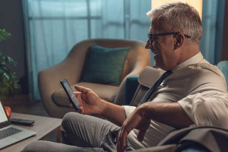 Corporate businessman relaxing on the sofa at home, he is smiling and connecting online using his...
