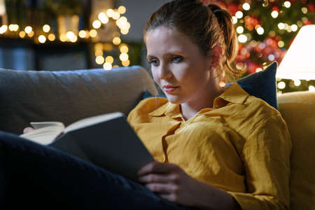 Happy young woman lying on the sofa at home and reading a book during Christmas holidaysの写真素材