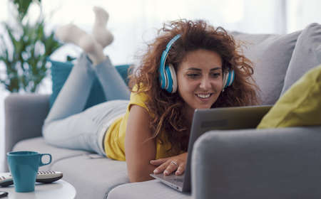 Happy woman relaxing at home, she is lying belly down on the sofa and watching videos on her laptop