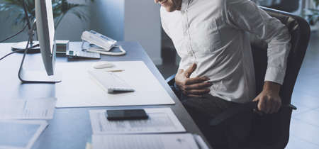 Businessman sitting at office desk and having stomach ache, he is touching his belly with his handの写真素材