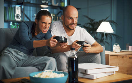 Couple sitting on the couch at home and playing video games together, they are holding the...