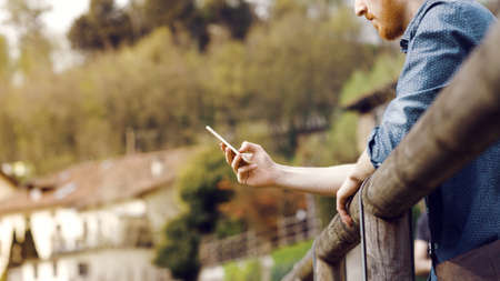 Hipster young man having a walk and texting with his mobile phone, he is leaning on a wooden railingの写真素材