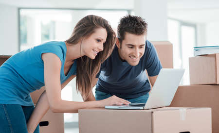 Happy couple connecting to the internet using a laptop in their new house surrounded by carton boxes