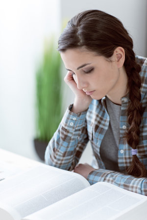 Bored young student girl sitting at desk in the library and reading a book, she is studying and leaning on her handの写真素材