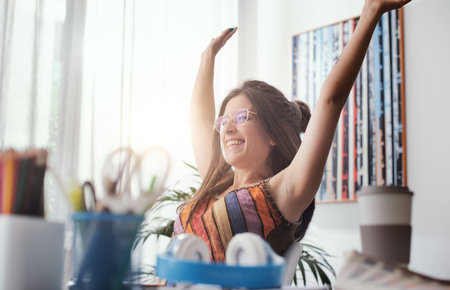 Cheerful successful woman celebrating with arms raised, she is sitting at desk at home and connecting with her computerの写真素材