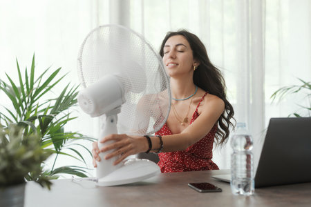Woman sitting at desk and cooling herself with an electric fanの写真素材