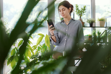 Woman in a room full of plants, she is connecting with her smartphoneの写真素材