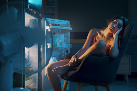 Exhausted woman suffering from the heat during the summer heatwave, she is sitting in front of the open fridge and cooling herselfの写真素材