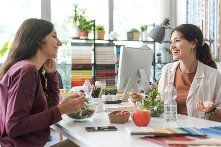 Women having a lunch break together in the office, they are eating salad and talking to each otherの写真素材