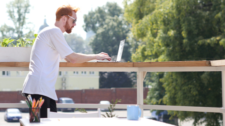 Young Man Working on Laptop, Typing, Standing in Balcony Outdoorの写真素材