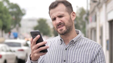 Outdoor Beard Casual Man Upset by Loss while Using Smartphoneの写真素材