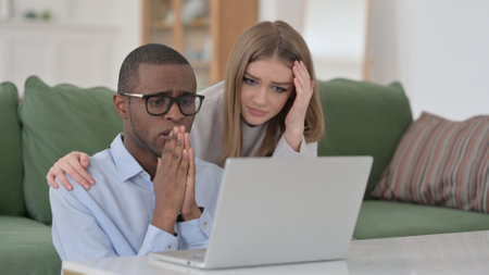 Interracial Couple Reacting to Loss while using Laptop at Homeの写真素材