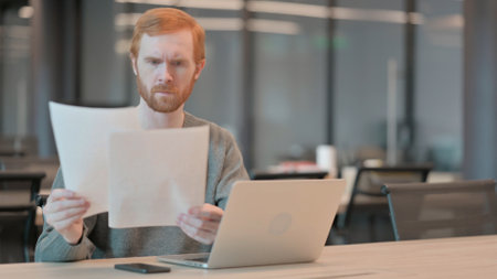 Young Man with Laptop Reading Documents in Officeの写真素材