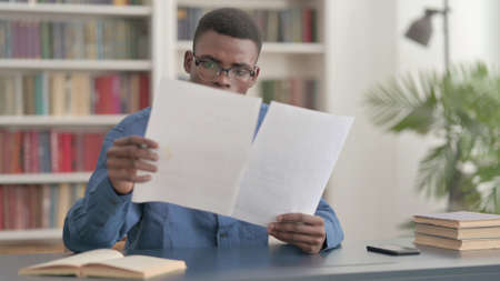 Young African Man Reading Reports while Sitting in Officeの写真素材