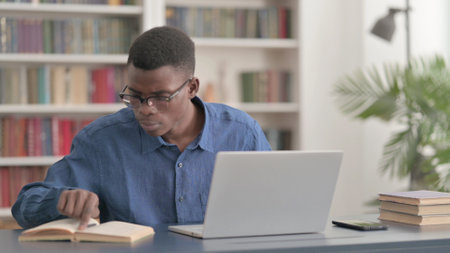 Young African Man Reading Book and Working on Laptop in Office