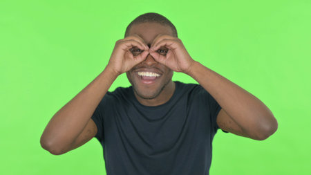 Searching Young African Man with Binocular Gesture on Green Backgroundの写真素材
