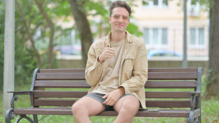 Thumbs Up by Casual Young Man Sitting Outdoorの写真素材