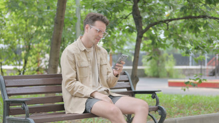Modern Young Man Facing Loss on Smartphone while Sitting on Benchの写真素材