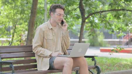 Modern Young Man Talking on Phone with Laptop in Lap Outdoorの写真素材