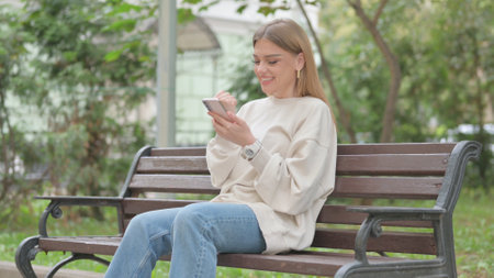 Casual Young Woman Celebrating Online Success on Phone while Sitting Outdoorの写真素材