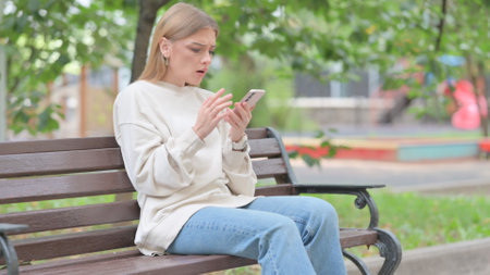 Young Woman Shocked by Loss on Phone while Sitting Outdoor on a Benchの写真素材