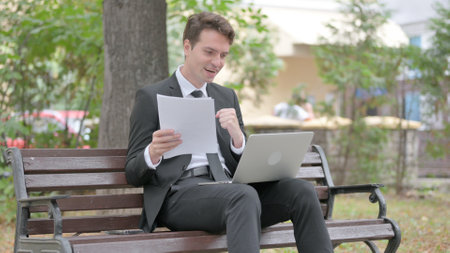 Young Businessman Celebrating while Using Laptop and Documents Outdoorの写真素材