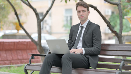 Businessman Looking at Camera with Laptop in Lap Outdoorの写真素材