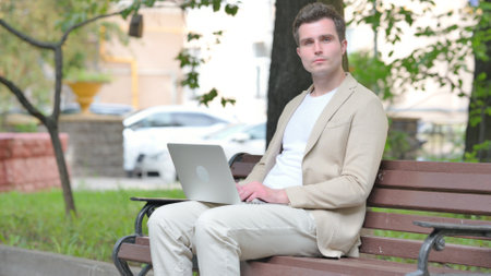 Casual Young Man Looking at Camera while Working on Laptop Outdoorの写真素材