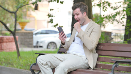 Casual Young Man Celebrating Success on Smartphone while Sitting Outdoor on a Benchの写真素材