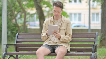 Casual Young Man using Tablet while Sitting Outdoor on a Benchの写真素材