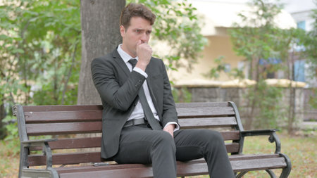 Coughing Young Businessman Feeling Sick while Sitting Outdoorの写真素材