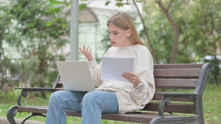 Casual Young Woman Feeling Upset while Reading Contract and Using Laptop Outdoorの写真素材