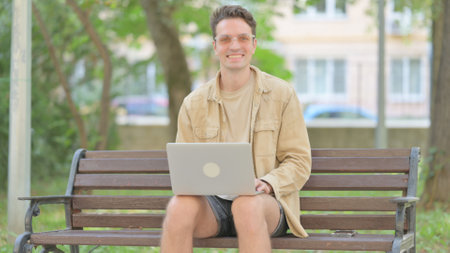 Casual Young Man Smiling at Camera while Working on Laptop Outdoorの写真素材