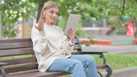 Young Woman Doing Video Chat on Tablet while Sitting Outdoor on a Benchの写真素材