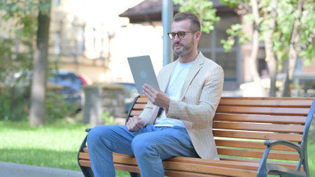 Middle Aged Man Doing Video Chat on Tablet while Sitting Outdoor on a Benchの写真素材