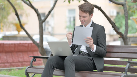 Businessman celebrating while using a laptop and documents outdoors.の写真素材