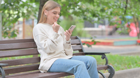 Young Woman Celebrating Online Success on Phone while Sitting Outdoorの写真素材