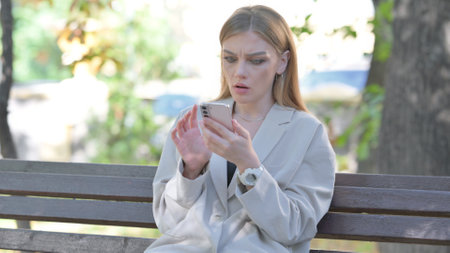 Young Businesswoman Shocked by Loss on Phone while Sitting on Bench on a Benchの写真素材