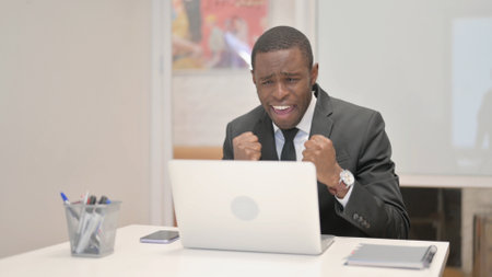 Excited African Businessman Celebrating Success on Laptop in Officeの写真素材